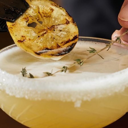 Bee’s Knees cocktail in coupeglas met suikerrand, gegarneerd met gegrilde citroen en tijm – klassieke roaring twenties cocktail.